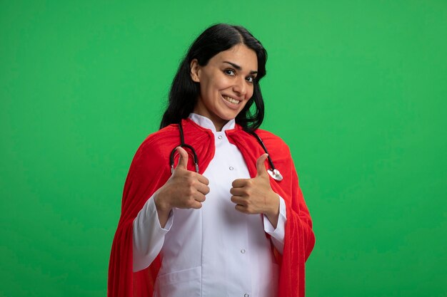 Sonriente joven superhéroe vestida con bata médica con estetoscopio mostrando los pulgares para arriba aislados en verde