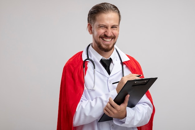 Sonriente joven superhéroe vistiendo bata médica con estetoscopio sosteniendo el portapapeles con lápiz aislado sobre fondo blanco.