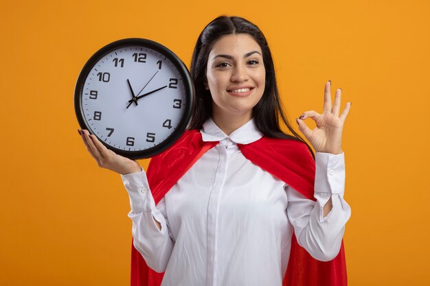 Sonriente joven superwoman sosteniendo el reloj mirando al frente haciendo bien signo aislado en la pared naranja
