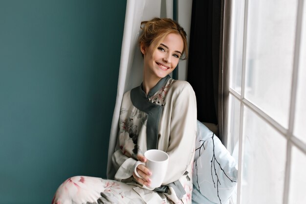 Sonriente joven con una taza de café, té en ella, sentada en el alféizar de la ventana, buenos días relajarse. Viste pijama de seda con flores, tiene cabello rubio. Foto en colores turquesa.