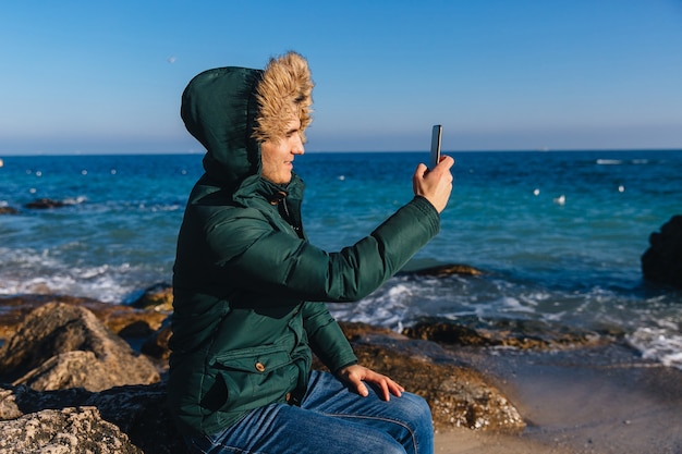 Sonriente joven tomando un selfie en teléfono celular cerca del mar. Vestido con una cálida chaqueta con pelo