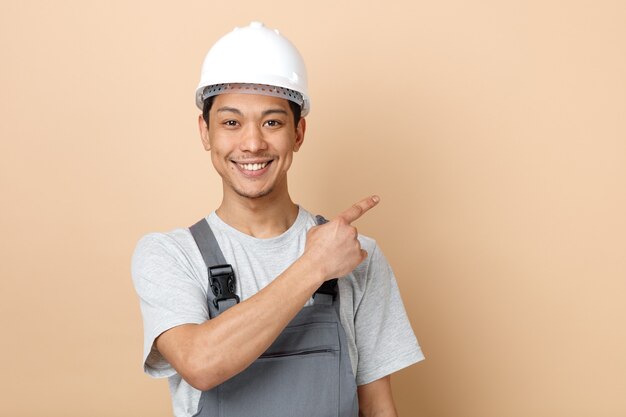 Sonriente joven trabajador de la construcción con casco de seguridad y uniforme apuntando hacia arriba en la esquina