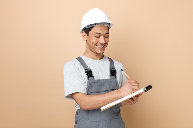 Sonriente joven trabajador de la construcción con casco de seguridad y uniforme escribiendo con lápiz en el bloc de notas