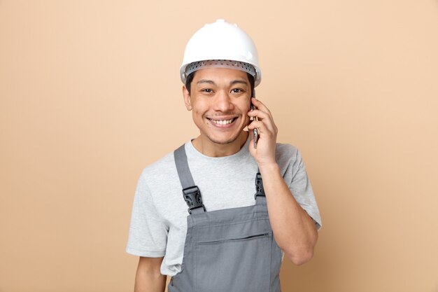 Sonriente joven trabajador de la construcción con casco de seguridad y uniforme hablando por teléfono