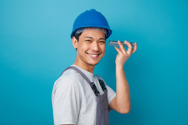 Sonriente joven trabajador de la construcción con casco de seguridad y uniforme de pie en la vista de perfil sosteniendo el teléfono móvil cerca de la oreja