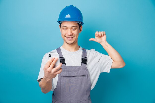 Sonriente joven trabajador de la construcción con casco de seguridad y uniforme sosteniendo y mirando el teléfono móvil apuntando a sí mismo