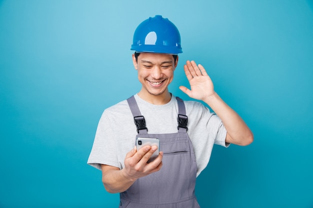 Sonriente joven trabajador de la construcción con casco de seguridad y uniforme sosteniendo y mirando el teléfono móvil ondeando