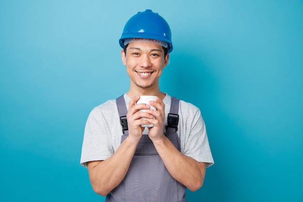 Sonriente joven trabajador de la construcción con casco de seguridad y uniforme sosteniendo una taza de té con ambas manos