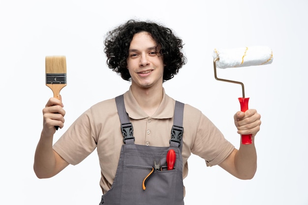 Sonriente joven trabajador de la construcción con uniforme sosteniendo pincel y rodillo de pintura mirando a la cámara con destornillador y alicates en el bolsillo aislado en fondo blanco