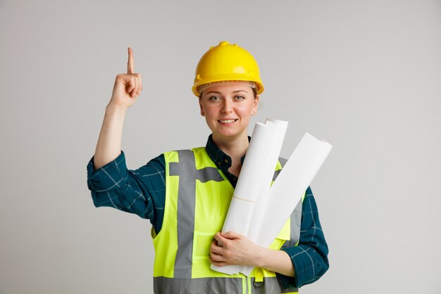 Sonriente joven trabajadora de la construcción con casco de seguridad y chaleco de seguridad sosteniendo papeles apuntando hacia arriba