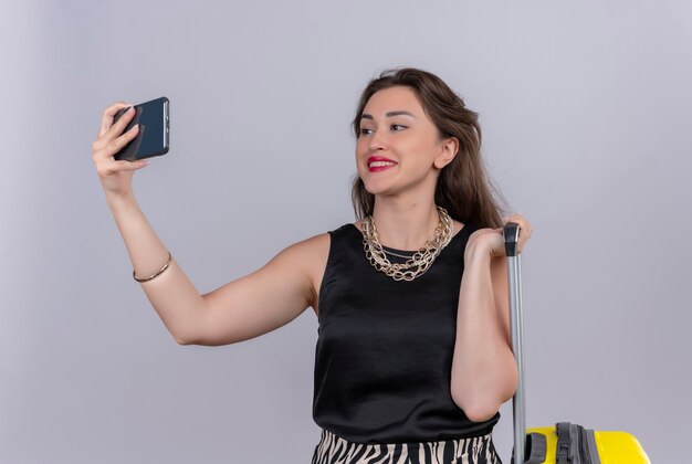 Sonriente joven viajera vistiendo camiseta negra con maleta y tomar un selfie en pared blanca