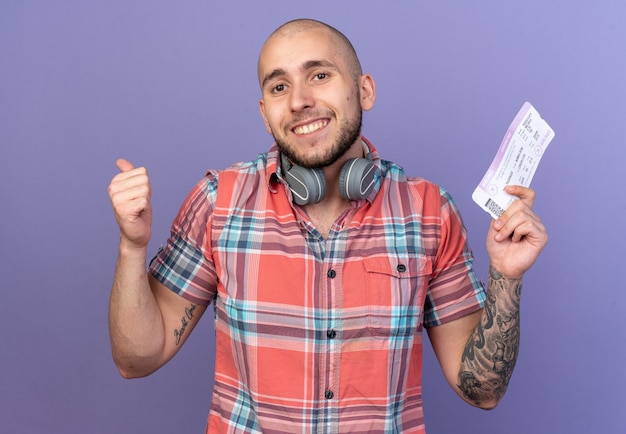 Sonriente joven viajero caucásico con auriculares alrededor de su cuello sosteniendo boleto aéreo y pulgar hacia arriba aislado sobre fondo púrpura con espacio de copia