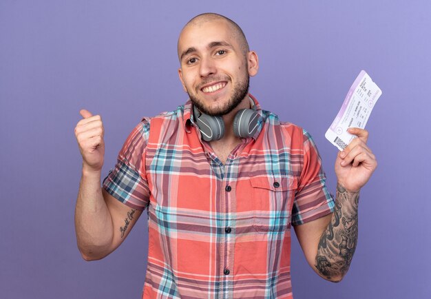 Sonriente joven viajero caucásico con auriculares alrededor de su cuello sosteniendo boleto aéreo y pulgar hacia arriba aislado sobre fondo púrpura con espacio de copia