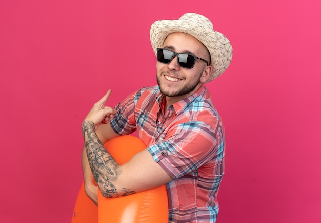 Sonriente joven viajero caucásico con sombrero de playa de paja en gafas de sol sosteniendo el anillo de natación y apuntando hacia atrás aislado en la pared rosa con espacio de copia