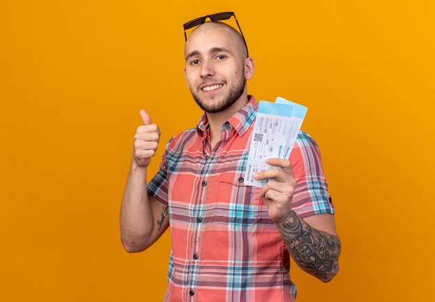 Sonriente joven viajero hombre con gafas de sol sosteniendo boletos de avión y pulgar hacia arriba aislado en la pared naranja con espacio de copia