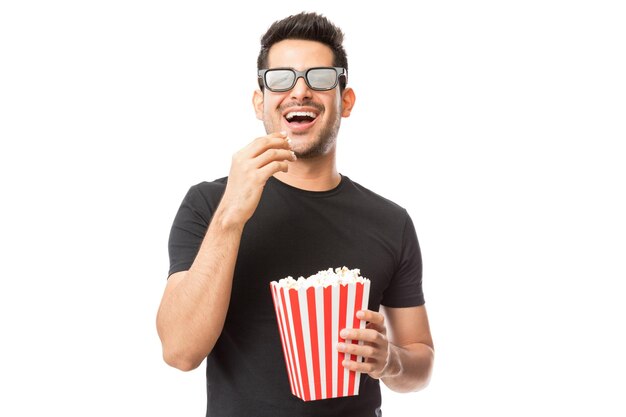 Sonriente joven viendo películas en 3D mientras come palomitas de maíz sobre fondo blanco.