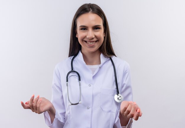 Sonriente joven vistiendo bata médica estetoscopio extendiendo las manos a la cámara sobre fondo blanco aislado