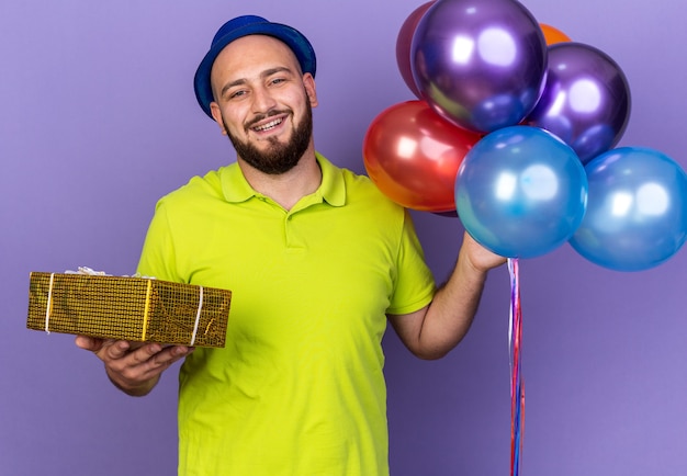 Sonriente joven vistiendo gorro de fiesta sosteniendo globos con caja de regalo