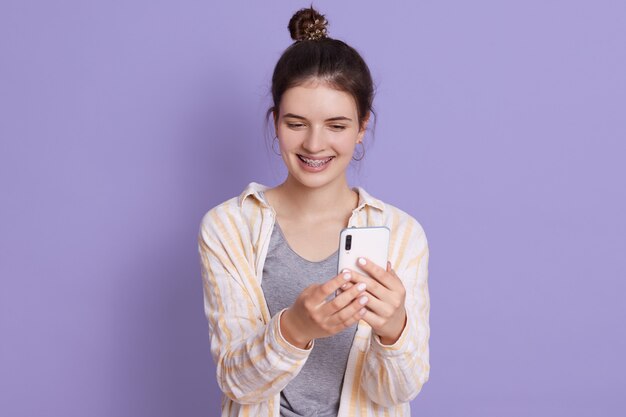 Sonriente jovencita con moño sosteniendo moderno teléfono inteligente en manos y haciendo selfie