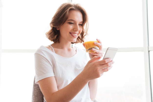 Sonriente jovencita sentada cerca de la ventana bebiendo jugo