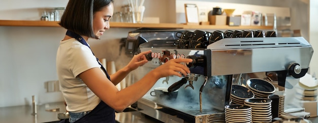 Sonriente linda chica coreana barista trabajando con la máquina de café preparar capuchino usando steamr en la cafetería