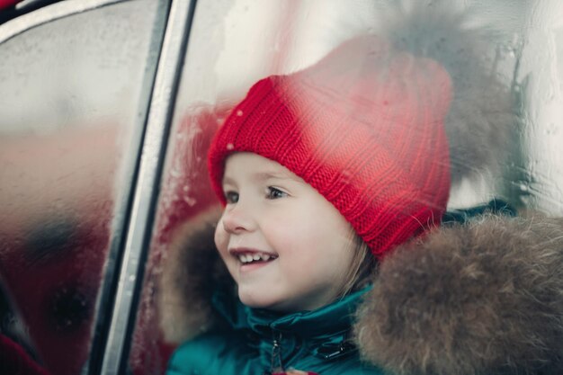 Sonriente linda chica de invierno con sombrero rojo sentado en el coche divirtiéndose en plano medio. Feliz hermosa bebé en ropa de abrigo con emoción positiva al aire libre rodeado de copos de nieve disfrutando de la infancia