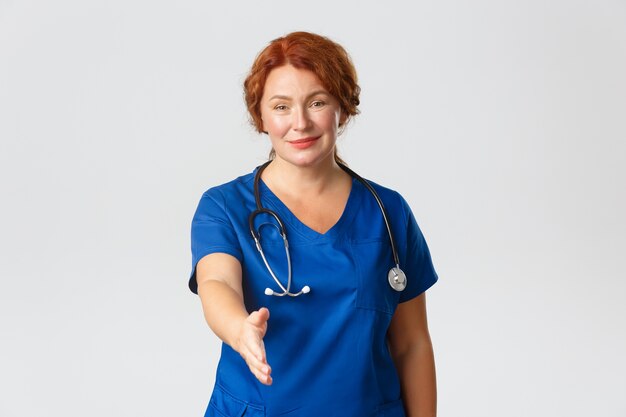 Sonriente y linda enfermera de mediana edad, doctora vestida de azul con aspecto amistoso, extender la mano para un apretón de manos, presentarse,