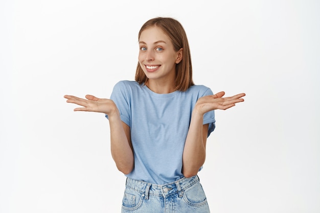 Sonriente linda mujer rubia encogiéndose de hombros, extendiendo las manos hacia los lados culpables, disculpándose, cometiendo un pequeño error, de pie tontamente contra la pared blanca.