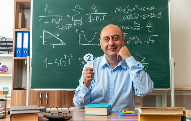 Sonriente maestro de mediana edad se sienta a la mesa con útiles escolares sosteniendo ventiladores de número en el aula