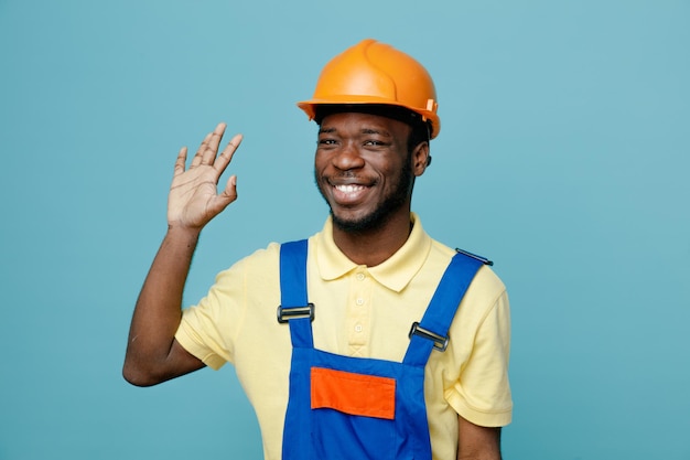 Sonriente mano levantada joven constructor afroamericano en uniforme aislado sobre fondo azul.