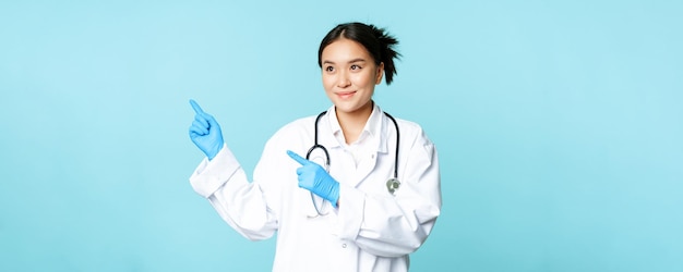 Sonriente médico asiático trabajador médico en uniforme señalando y mirando a la izquierda en el espacio de la copia de pie sobre fondo azul.