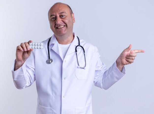 Sonriente médico varón de mediana edad vistiendo bata médica y un estetoscopio mostrando el paquete de tabletas mirando al frente apuntando al lado aislado en la pared blanca