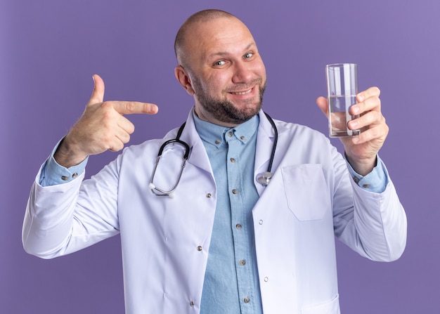 Sonriente médico varón de mediana edad vistiendo bata médica y estetoscopio sosteniendo y apuntando al vaso de agua aislado en la pared púrpura