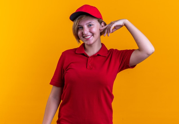 sonriente mirando al frente joven repartidora vistiendo uniforme con gorra aislado en la pared naranja