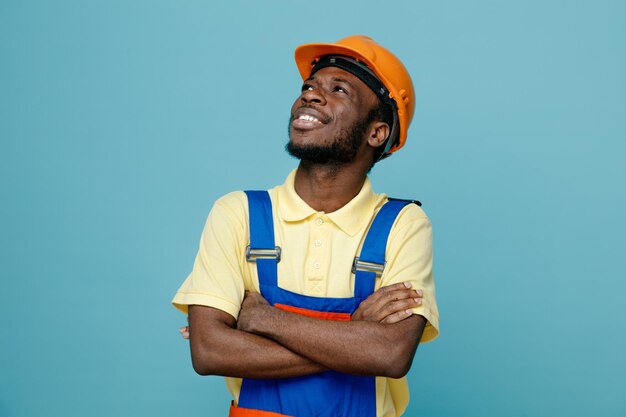 Sonriente mirando el lado cruzando las manos joven constructor afroamericano en uniforme aislado sobre fondo azul.