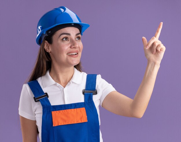 Sonriente mirando a la mujer joven constructor de lado en puntos uniformes en el lado aislado en la pared púrpura