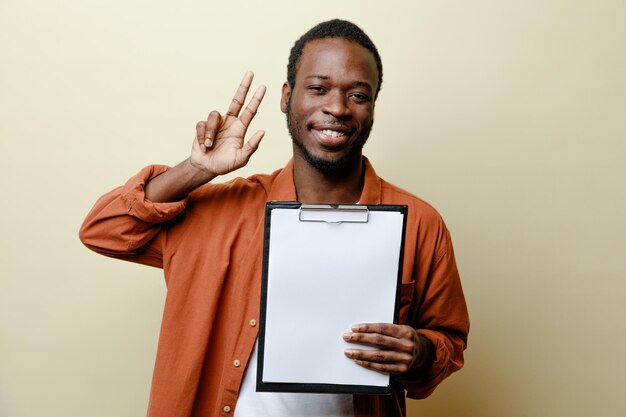 Sonriente mostrando gesto de paz joven afroamericano sosteniendo portapapeles aislado sobre fondo blanco.