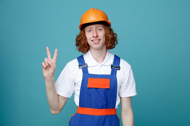 Sonriente mostrando número joven constructor hombre en uniforme aislado sobre fondo azul.