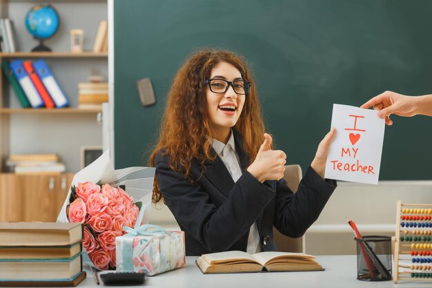 sonriente mostrando los pulgares hacia arriba la joven maestra recibió una postal sentada en el escritorio con herramientas escolares en el aula