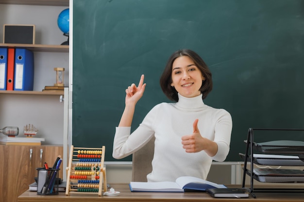 sonriente mostrando los pulgares hacia arriba joven maestra sentada en el escritorio con herramientas escolares en el aula