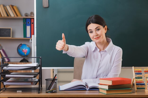 sonriente mostrando los pulgares hacia arriba joven maestra sentada en el escritorio con herramientas escolares en el aula