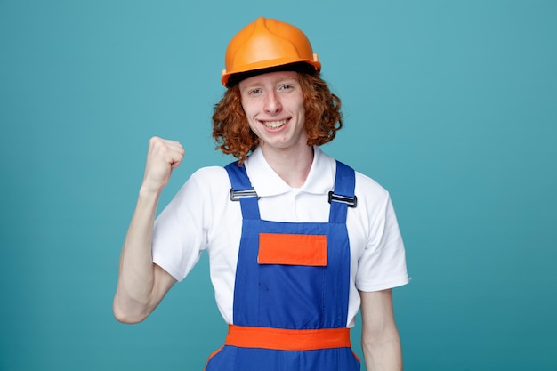 Sonriente mostrando puño joven constructor hombre en uniforme aislado sobre fondo azul.