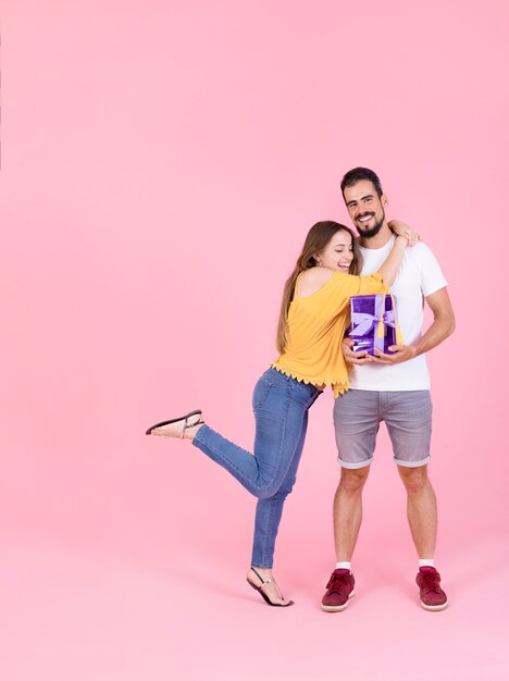 Sonriente mujer abrazando a su novio con caja de regalo contra el fondo rosa