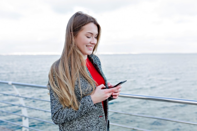 Sonriente mujer adorable en abrigo gris y camisa roja smartphone de desplazamiento en el terraplén y disfruta del buen tiempo