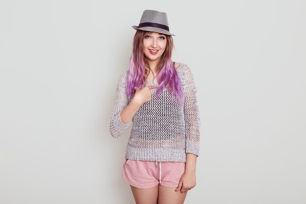 Sonriente mujer adorable con sombrero y apuntando a sí misma con el dedo índice, vestida con camisa y elegante sombrero, siendo increíble que la elijan, aislada sobre una pared blanca.