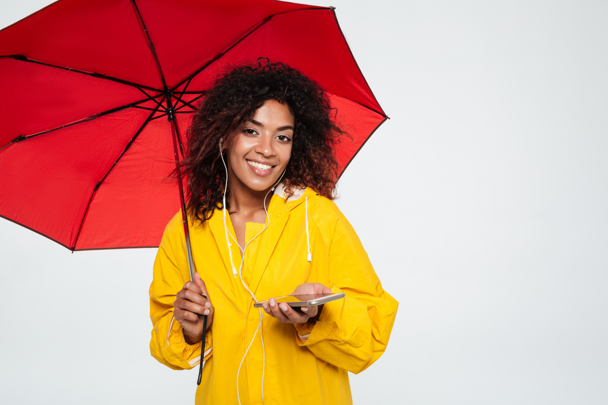Sonriente mujer africana en gabardina escondiéndose bajo el paraguas y escuchando música