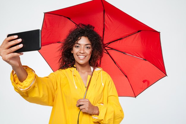 Sonriente mujer africana en gabardina escondiéndose bajo el paraguas y haciendo selfie en su teléfono inteligente sobre fondo blanco.