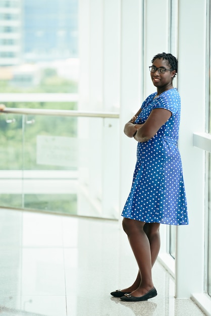 Sonriente mujer africana en vestido azul de lunares apoyada contra el marco de la ventana y sonriendo