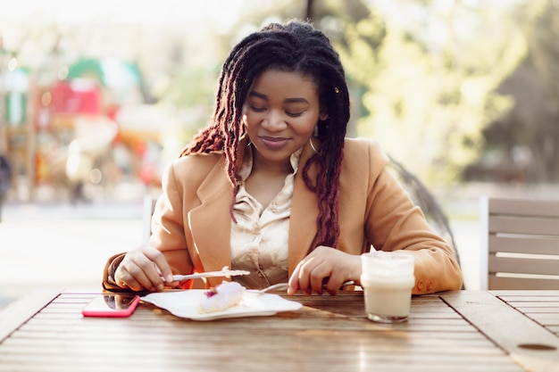 Sonriente mujer afroamericana bebiendo café y comiendo postre en la cafetería al aire libre