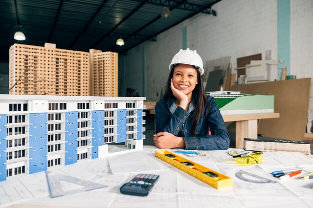 Sonriente mujer afroamericana en casco de seguridad cerca del modelo de edificio